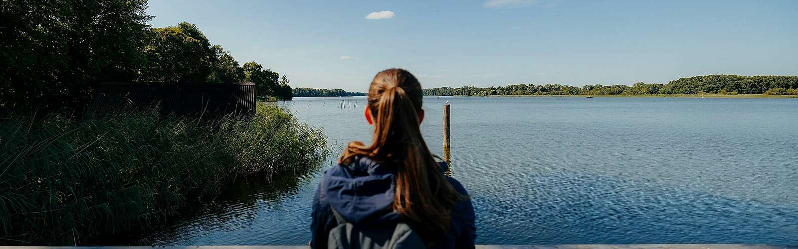 Eine junge Frau steht an einem Geländer und schaut über den blauen See. Sie ist von hinten zu sehen.,
            
        
                Foto: Tourismusverband Ruppiner Seenland e.V./Julia Nimke